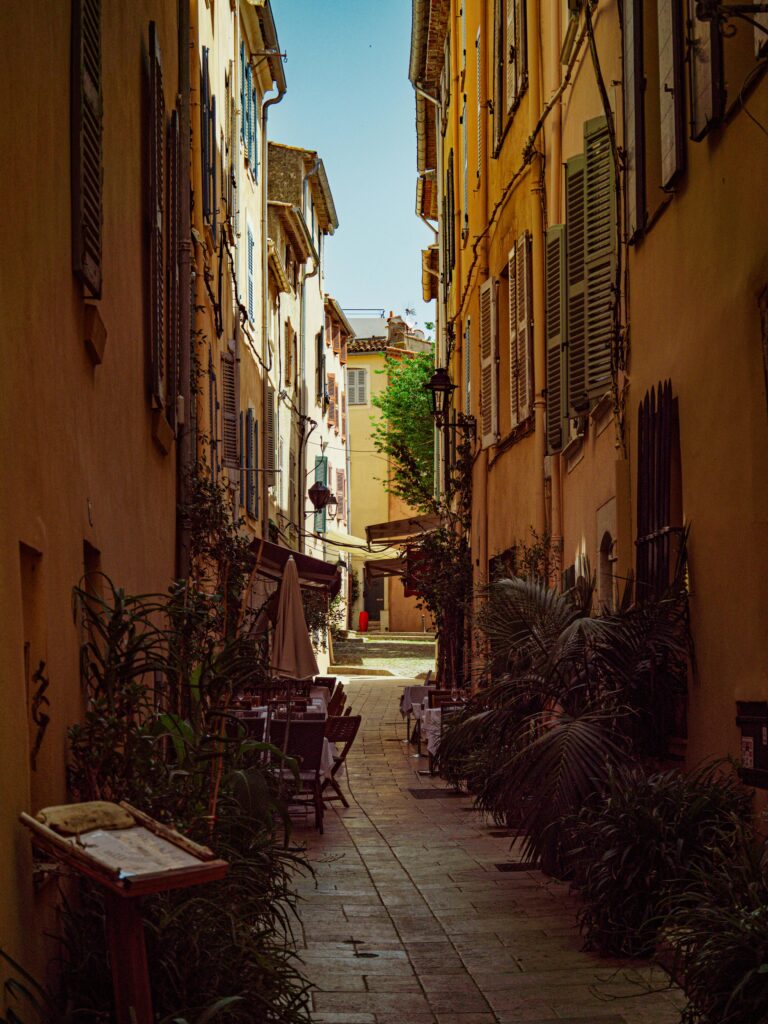Explore a quaint Mediterranean alley with charming outdoor café seating, bathed in warm sunlight.