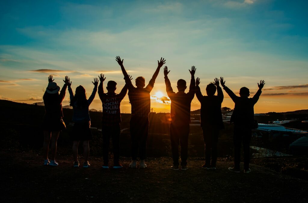 A diverse group of friends raises their arms in celebration against a vibrant sunset backdrop. مرفأ الديوانية